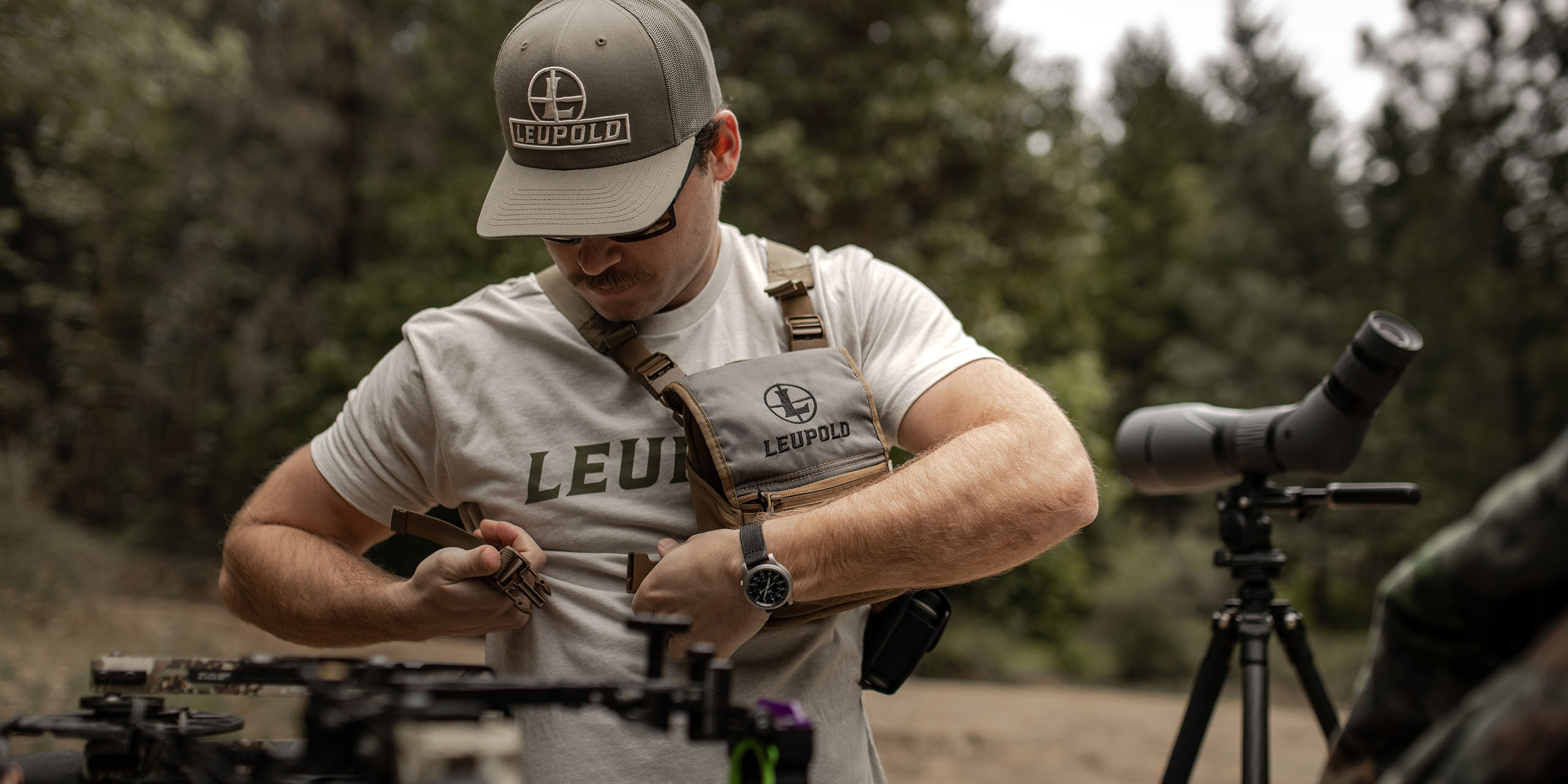 Male model getting outfitted for a hunt. They are adjusting their Leupold Binocular Harness System while wearing a Leupold hat and T-Shirt. 