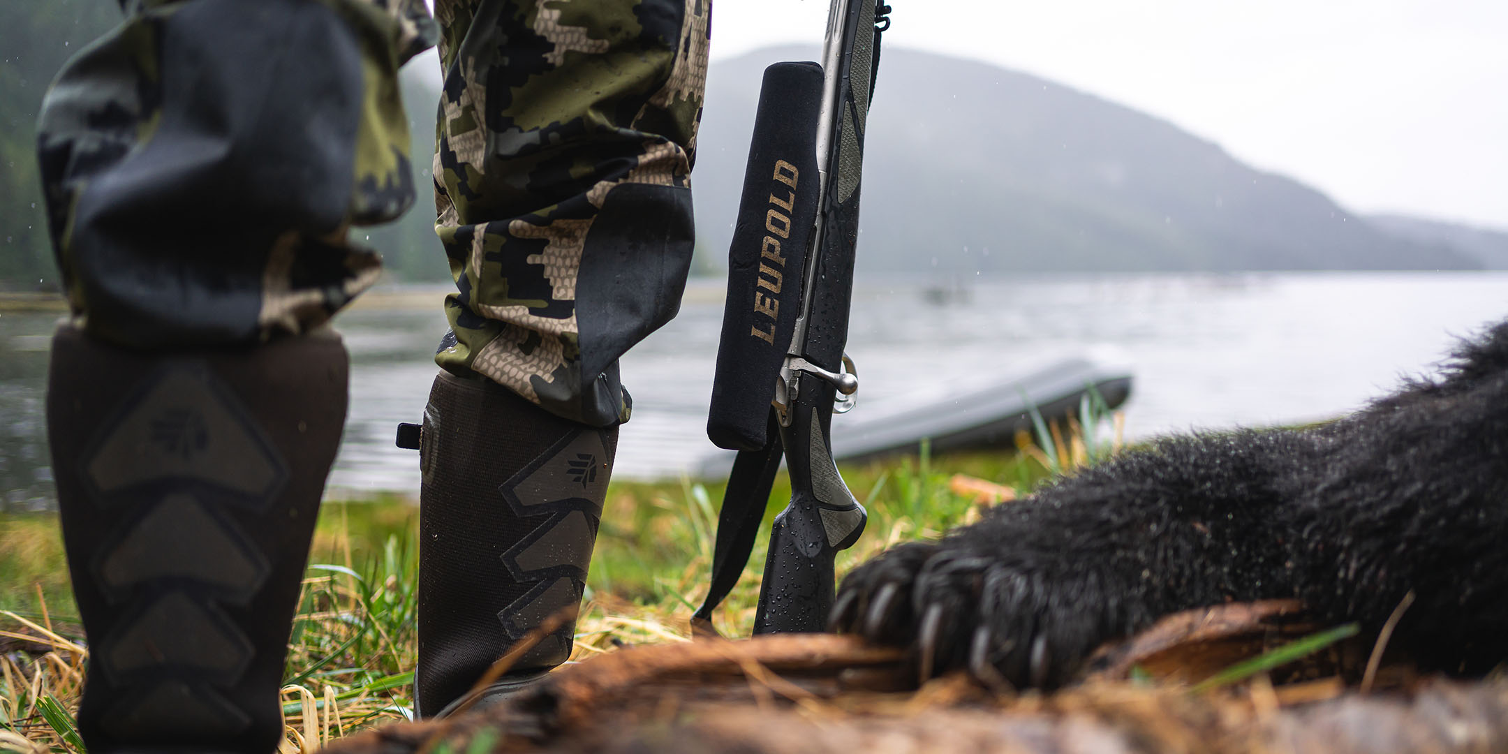 A bear paw outstretched on a log. Nearby a person's legs are visible with their rifle and Leupold riflescope next to them.