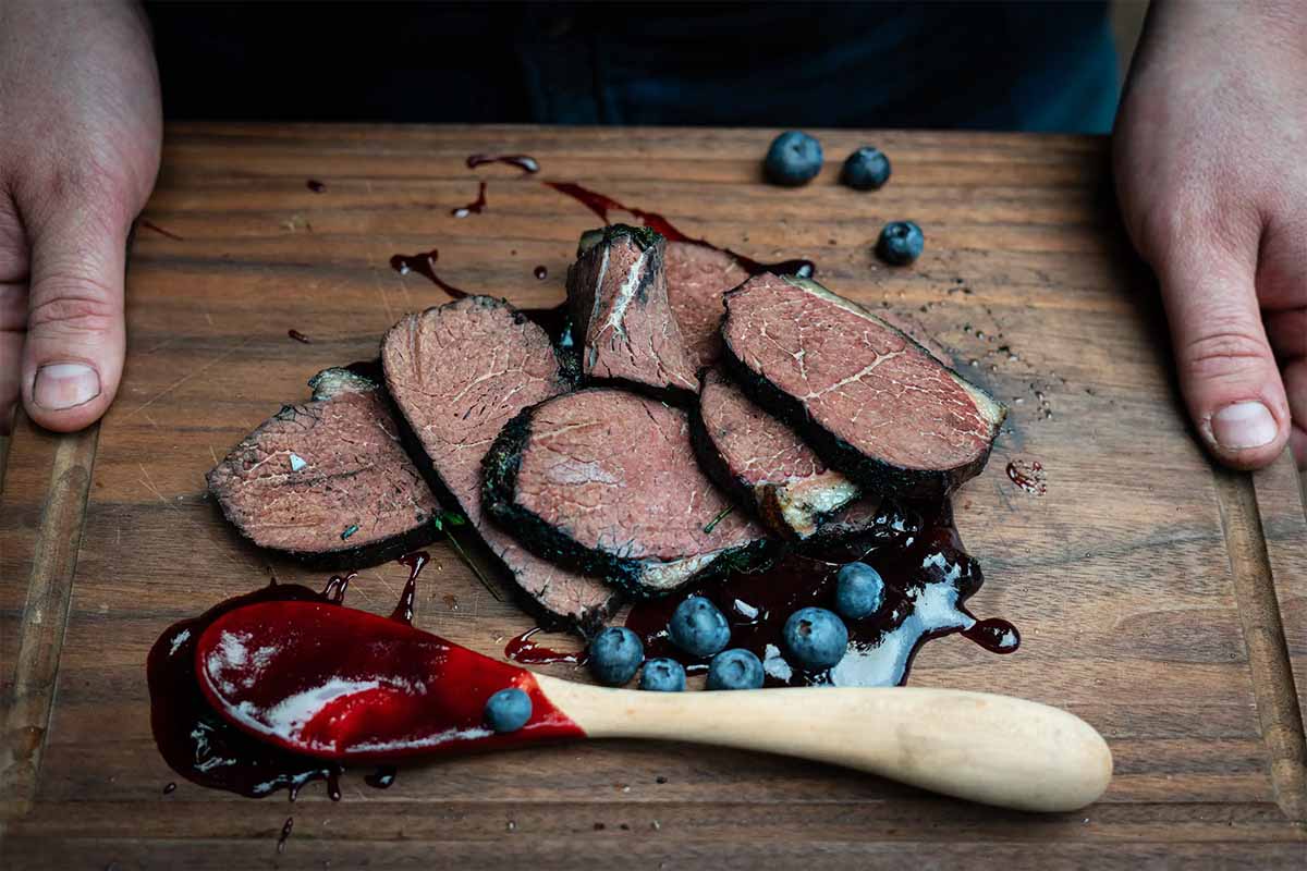 A cutting board with cuts of bear meat , blueberries, and blueberry compote. Lead image for "Field to Table: Bourbon Blueberry Bear Roast"