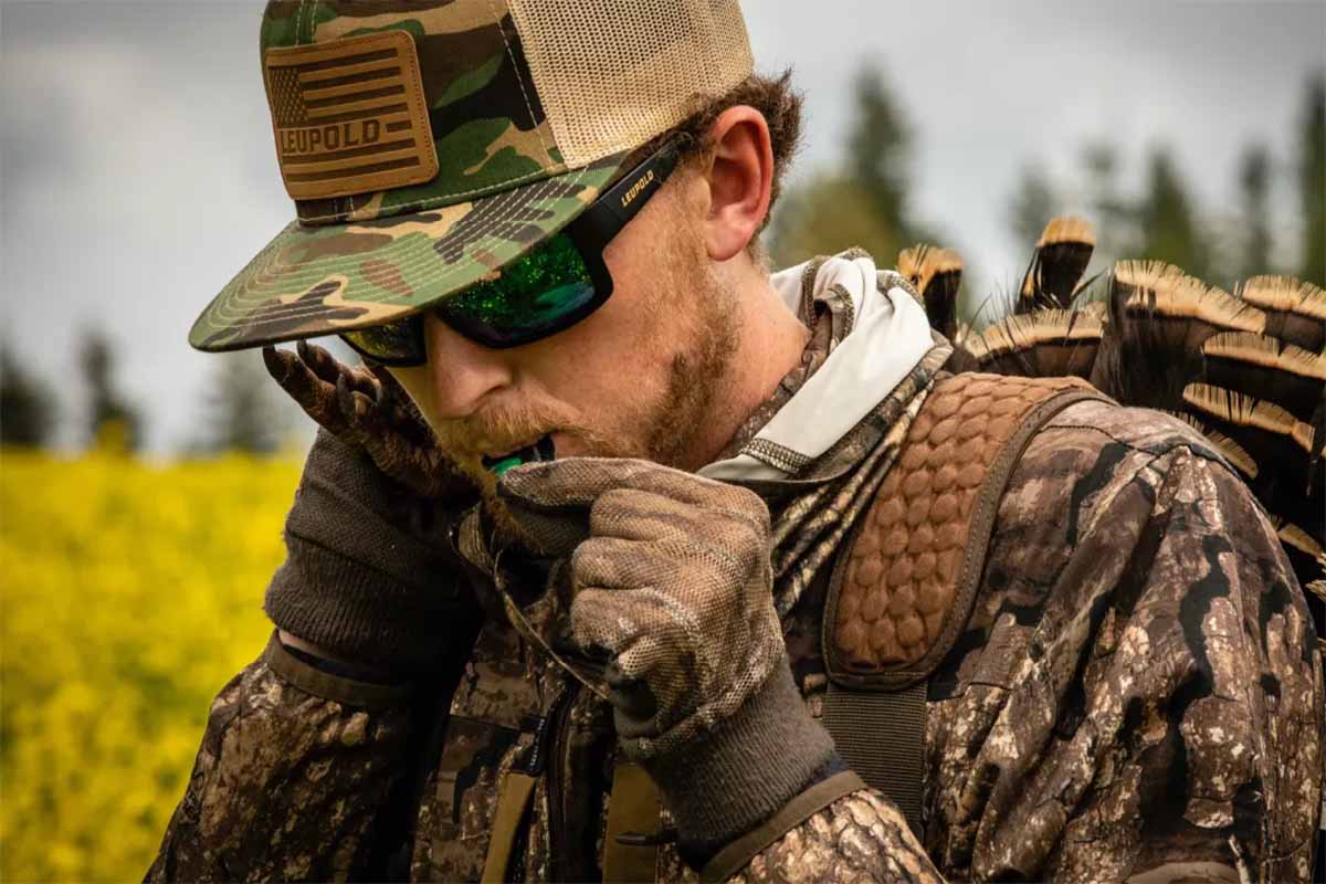 Upclose landscape image of a hunter using turkey caller adroned in Leupold hat, performance eyewear, and camo hunting gear.